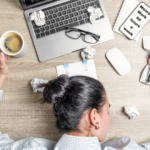 Overworked woman with head on desk, surrounded by crumpled paper, laptop, coffee, and a note that reads "Memo: Work 60 hours a week."