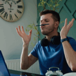 A man in a blue shirt balancing a pen on his upper lip while sitting at a desk with a laptop, headphones around his neck, and arms raised in frustration or boredom.