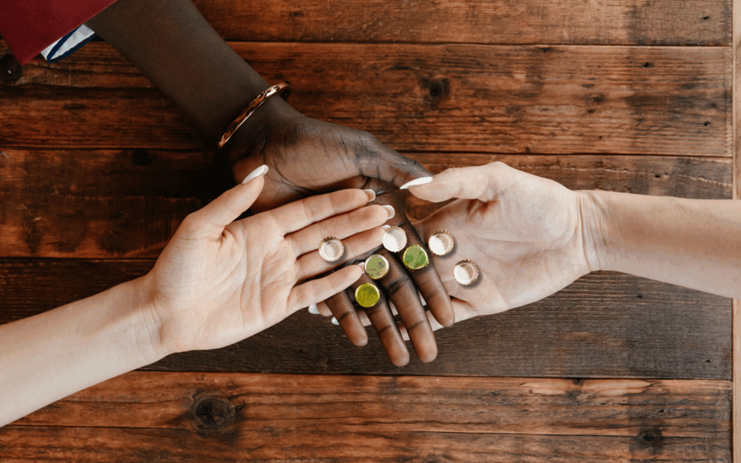 Three people of different skin tones holding bottle caps together on a wooden table, symbolizing unity and connection.